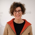 Headshot of artist Stephanie Rothenberg, wearing a tan cardigan with orange collar, and a black shirt, and black glasses. 