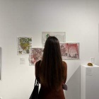 Back of the head/torso of Aidelen Montyoa, looking at an art gallery wall displaying her work of maps of Buffalo, red red-lining embroidery. 