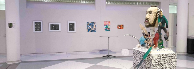 White walls with artwork hung from wire cables, and sitting on a pedestal. Gray and white checkered floor, in the UB Center for the Arts Atrium. 