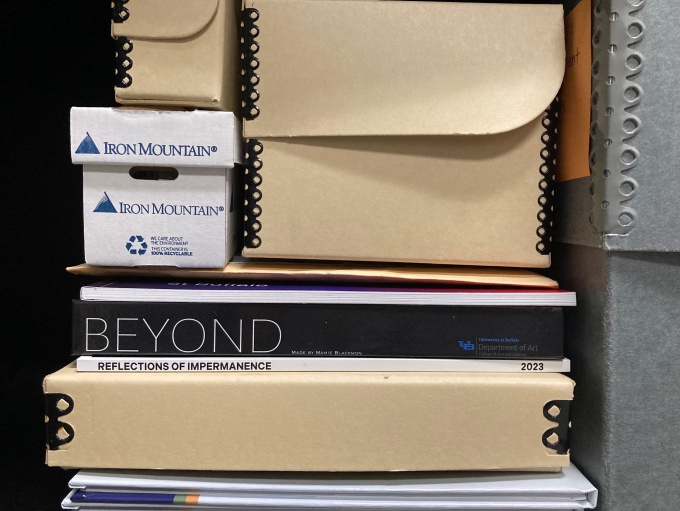 Closeup image of archival boxes if various sizes stacked with paperback exhibition catalogues on a shelf. 