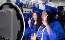 2 students posing in front of a photo booth wearing caps and gowns. 
