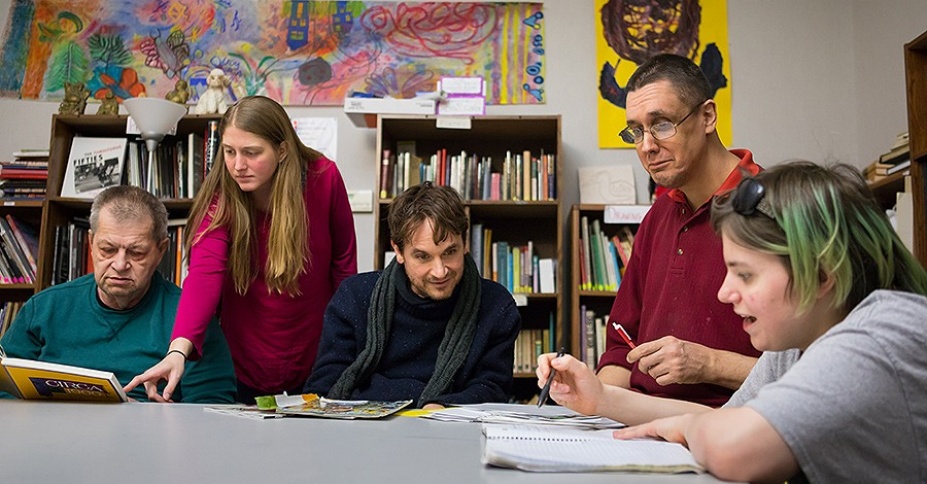 Pictured, five white-presenting individuals of various ages and genders – with Center for Disability Studies Director Michael Rembis in the center – sit at a table with books and papers before them, engaged in discussion. Behind them, the walls are lined with bookcases and covered with colorful art. 