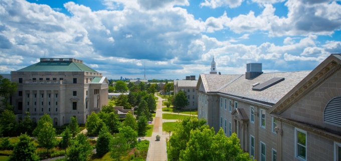 Pictured, an aerial view of several tan-colored stately stone buildings from UB’s South Campus. In the center of the frame, a sidewalk cuts vertically between the buildings and fades into the distance, with lush green trees and grass on either side and a bright blue sky with billowing white-grey clouds above. 