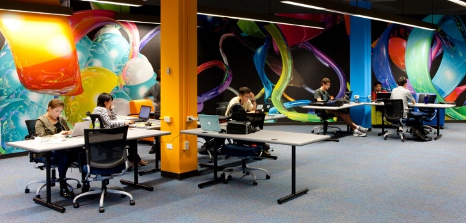 A small group of students work on laptops at desks arranged around colorful beams in a large study area with bright walls and fluorescent lights. 