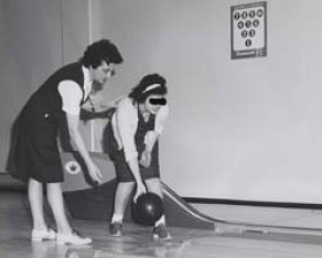 A historical black and white photo depicts a young white- and female-presenting resident about to release a bowling bowl onto the lane while a female staff member assists her. Black marker covers the resident’s eyes to maintain her privacy. 