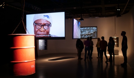 View of Lewuga Benson's installation of "The Land Gives Until It No Longer Can" which shows an oil drum hanging in the foreground, three hanging screens, one with an close-up image of a Black man wearing a winter cap and glasses. Several visitors are standing towards the back of the room. 