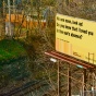 A billboard on a sunny winter day, with leafless trees and some vegetation. Train tracks intersect the photograph. The billboard, mostly in yellow, features a haiku, credited to Jason Livingston and Phoebe A. Cohen: “No one noon, look up! Do you know that I loved you in the sun’s absence?” Vertical lines in gradients of orange and yellow are next to the hashtag #InthePathofTotality, and Squeaky Wheel’s logo. 
