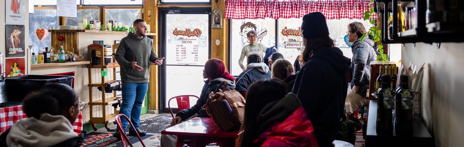 Students gather inside a small local restaurant during a class field trip, listening to a speaker near the entrance while seated at red tables.