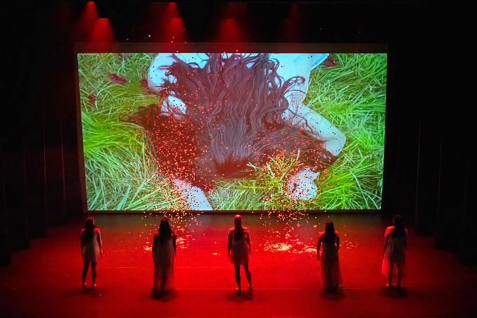 5 female dancers on stage face a movie screen with a young woman lying in green grass. 