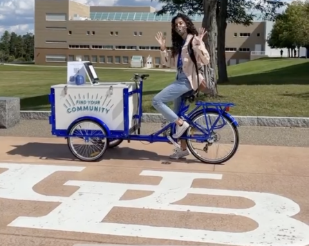 A student is waving while sitting on the Career Design Center's blue and white tricycle outside Clemens Hall.