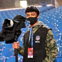 A student stands in front of empty football stadium seats with a camera in one hand. 
