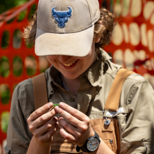 A person wearing a beige cap with a blue bull logo smiles while examining a small green object. They are dressed in overalls and a watch, with an orange safety net blurred in the background. The setting appears to be outdoors, possibly at a work or project site. The person's face is mostly obscured by the cap, but their body language conveys a sense of focus and happiness. 