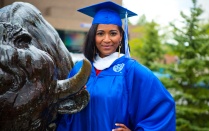 A student stands by the Buffalo statue in graduation robes. 