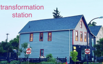 Light blue house with red trimmed windows, on a corner of a street with a fire hydrant in frotn and pine tree behind. 