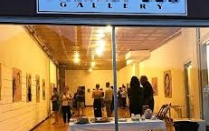 View of a long skinny art gallery through the street level front windows, with over a dozen people viewing artwokr hung on the walls, and the gallery name, Project 308, written on a sign above the window. 