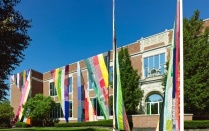 Brick building (UB Anderson Gallery), draped with patchwork tapestries, the height of the building, with trees and shrubs visible in the foreground and edges. 
