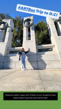 A young woman standing in front of a memorial with bronze wreths hanging from stone towers. 