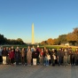 A group of thirty people, arranged in a two row line, pose on a gravel walkway at the National Mall in Wadshington D.C., with the Washington Monument visible in the background. It is close to sunrise, and the people shield their eyes from sunbeams coming across their faces. Grassy rear. 