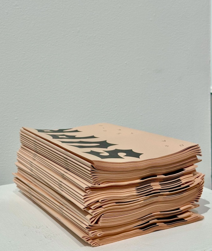 Zoom image: Stack of salmon pink newsprint paper catalogues for the art exhibition "Supreme", on a short white pedestal.