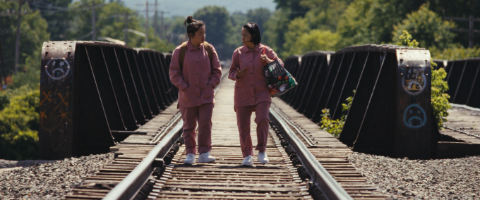 Still from the film "The Motherload", co-written and directed by Van Tran Nguyen. Image shows two women in pink jumpshuits walking between train tracks, having just crossed a bridge. Trees and hazy blue sky in background. 