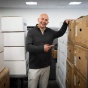 A man in a black sweater and white pants stands amidst stacks of cardboard boxes as tall as him, pointing at some, in an office with whtie tile drop ceilings. 