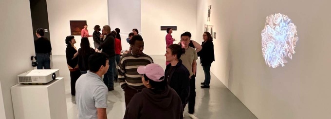 A crowd of people in a white walls and floors art gallery, with a projection on the right wall, and paintings hanging in the back. 