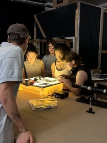 Zoom image: Photographic film negatives are illuminated on a small light-box work table. Overhead lights are turned off. Five people (college students) stand around the table and observe.
