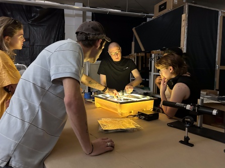 Zoom image: Artist John Opera points to photographic prints he is working on, on a small light-box work table. Overhead lights are turned off. Three people (college students) stand around the table and observe.