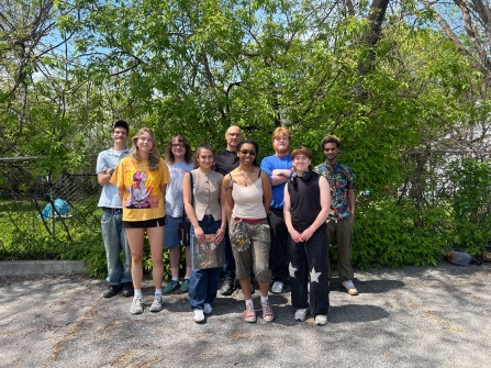 Nine people standing in two rows for a group portrait photo. Trees are in the background with blue skies peaking through, and black pavement underfoot. 