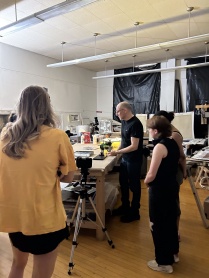 Zoom image: Artist John Opera points to photographic prints he is working on, on a large studio work desk in a high-ceilinged room with fluroescent drop lights. Three people (college students) stand around the table and observe. Windows in the background are covered with black plastic tarps.