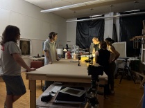 Zoom image: Artist John Opera points to photo films on a small light box, on top of a large studio work table. Overhead lights are turned off. Four people (college students) stand around the table and observe. Windows in the background are covered with black plastic tarps.