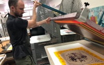 Noah Breuer lifts screenprint screen above a multicolo print. Paper drying racks in background. 