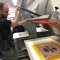 Noah Breuer lifts screenprint screen above a multicolo print. Paper drying racks in background. 