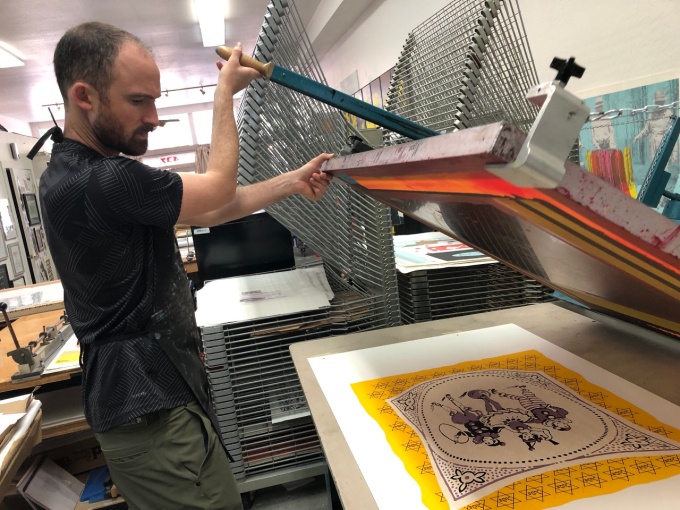 Noah Breuer lifts screenprint screen above a multicolo print. Paper drying racks in background. 