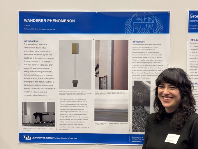 Zoom image: Ligia Sato, wearing black long-sleeved shirt and with sticker nametag on her chest, poses in front of a poster with columns of text and images related to her thesis project.