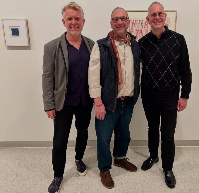 Three men standing in an art gallery, with two small framed works hanging behind. 