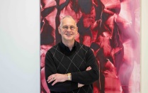 A man with short white and gray hair and glasses, wearing black button up shirt, standing in front of a shades of red and pink large abstract artwork. 