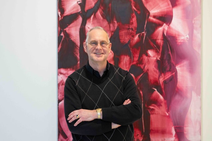 A man with short white and gray hair and glasses, wearing black button up shirt, standing in front of a shades of red and pink large abstract artwork. 