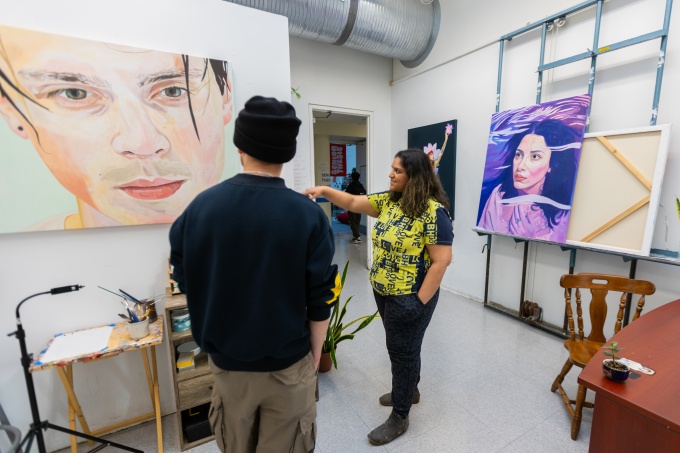 Two people standing looking at painting artwork mounted on white walls, in an art studio room, with an HVAC silver pipe above and open doorway at the rear. 