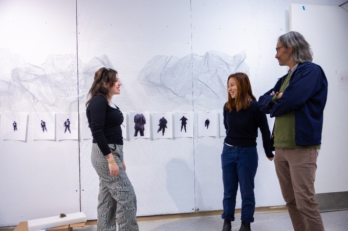 Three people standing in a white walled art studio room, with a wall of photographs of one person standing in various poses, pinned to the wall and wire clouds suspended above. 