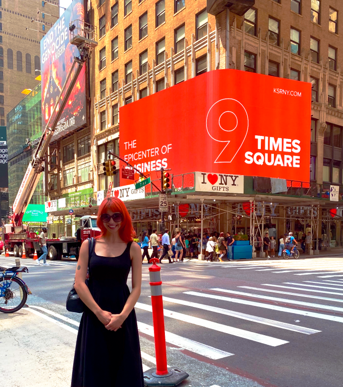 Woman stands in front of red background digital billboard in times square, attached to brick building. 