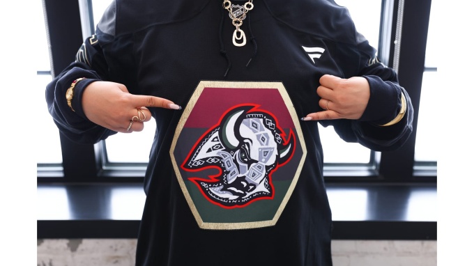 Zoom image: Closeup of black hockey practice jersey, featuring the Sabres Black History celebration crest designed by George Hughes. Person wearing the jersey points at the crest, with long, painted fingernails.