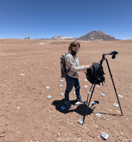A person standing by a tripod in a large bare dirt plain with rocky hills in the background. 