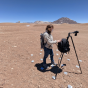 A person standing by a tripod in a large bare dirt plain with rocky hills in the background. 
