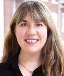 Headshot of a smiling person with long hair, bangs, wearing a black shirt and small necklace. 