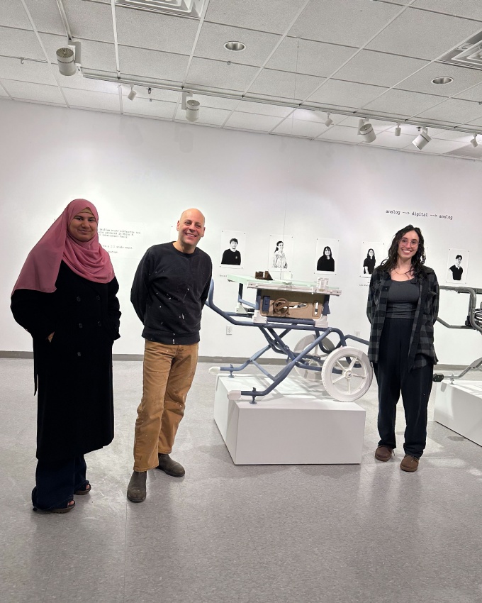 Three people stand in an art gallery with black and white artist photos on the far wall, and a replica tablesaw in the middle. 