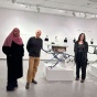 Three people stand in an art gallery with black and white artist photos on the far wall, and a replica tablesaw in the middle. 