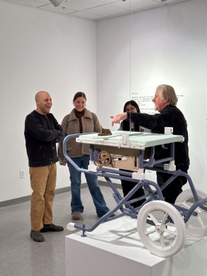 Four people stand in an art gallery with a replica tablesaw in the middle. 