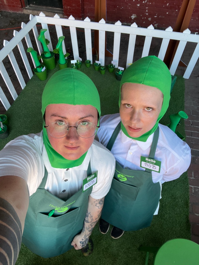 Downward angle selfie of two people wearing bright green fitted head covers, white shirts, and green aprons, standing in a paddock of fake grass with a white picket fence surrounding.