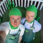Downward angle selfie of two people wearing bright green fitted head covers, white shirts, and green aprons, standing in a paddock of fake grass with a white picket fence surrounding. 